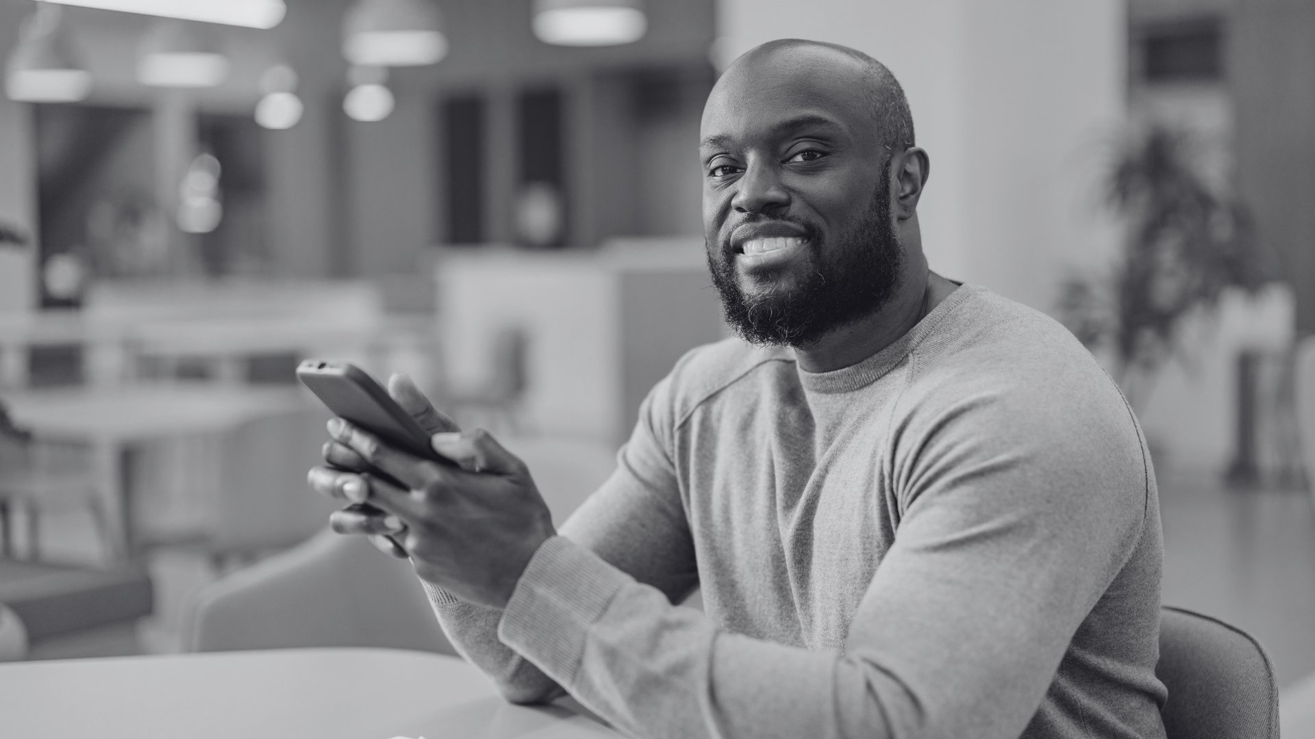 A black man smiling and looking at the camera while holding a cell phone. The phot is in black and white. The post is about Digital Skills and Digital Literacy.