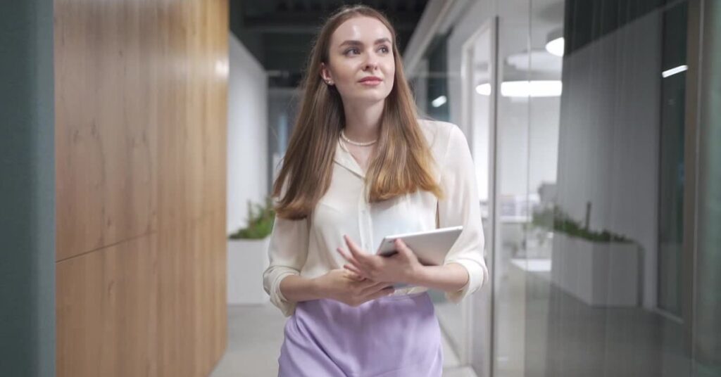 A confident young woman walks through an office, holding a tablet and showing calm focus—capturing the essence of quiet career confidence.