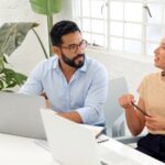 Two HR professionals having a collaborative discussion at a modern office desk with laptops open.