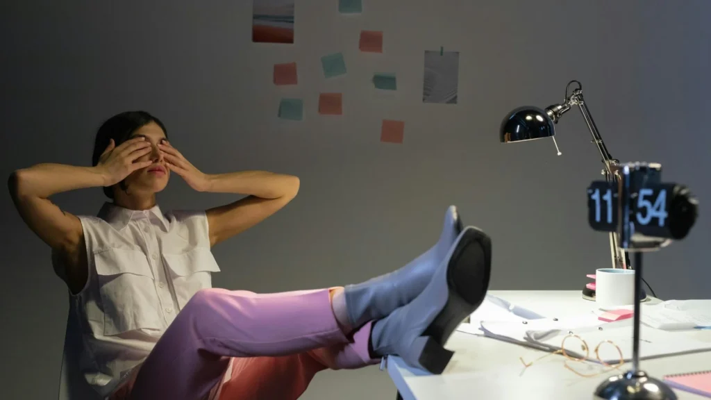 Woman reclining in an office chair with feet resting on a desk, hands covering the face, surrounded by a laptop, desk lamp, papers, and scattered sticky notes on the wall, illustrating workplace fatigue, overload, and signs of burnout in a demanding work environment.