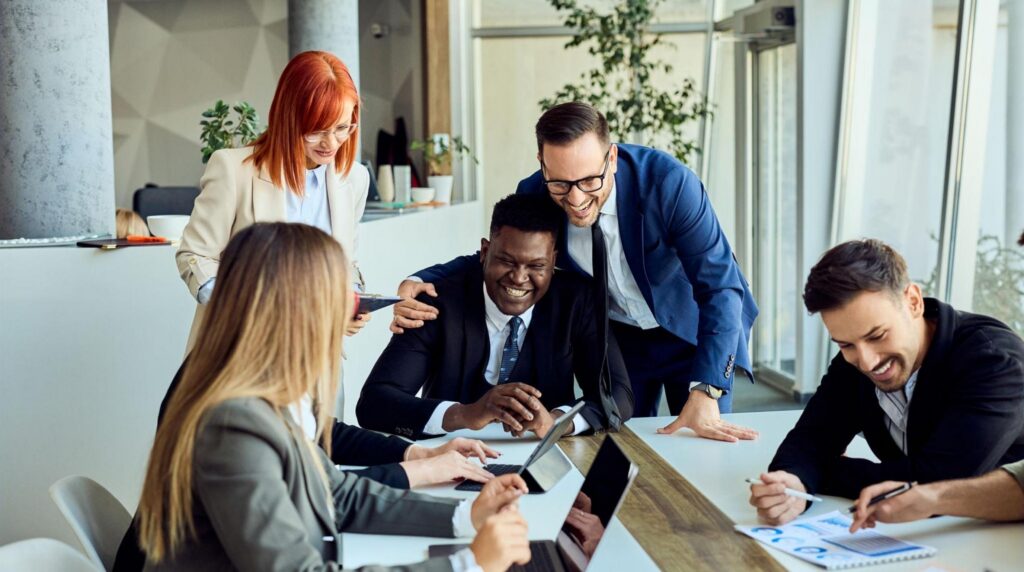 A group of professionals gathered around a table in a modern office collaborate on a project, reviewing documents and a tablet while engaging in discussion and celebarting the successfull onboarding of a new colleague.
